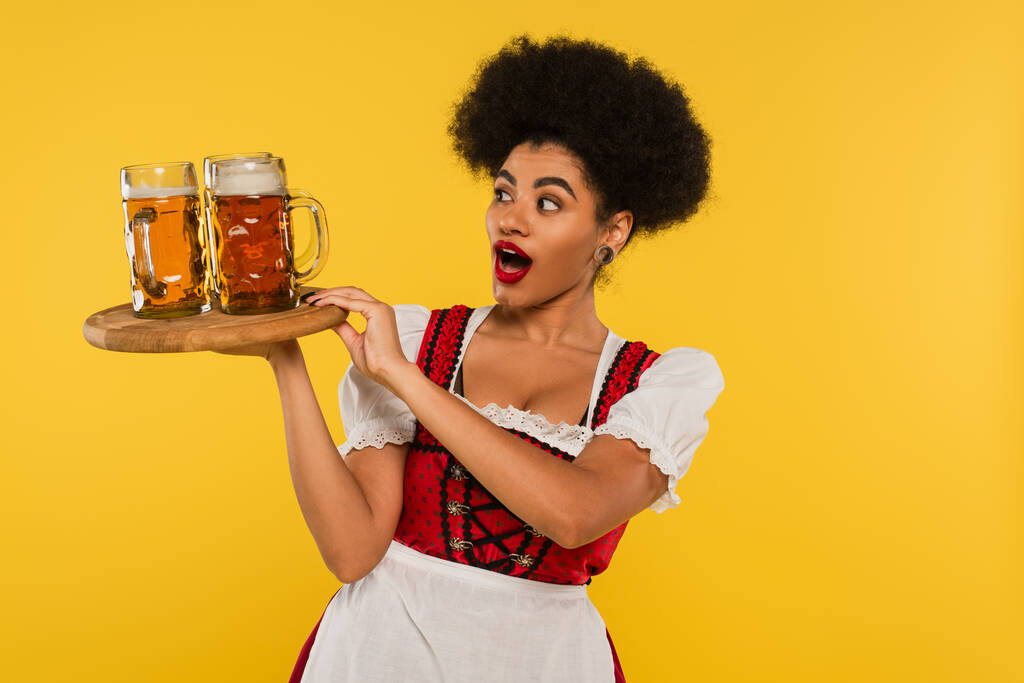 изумленная африканская официантка Октоберфест держит деревянный поднос с кружками пива на желтом, баннер - Фото, изображение