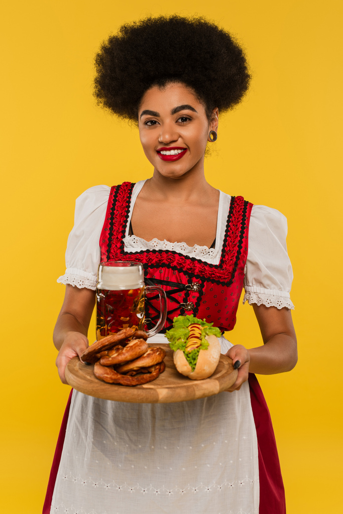 χαρούμενη αφρικάνα αμερικανίδα σερβιτόρα που κρατάει ξύλινο δίσκο με κουλούρια και χοτ ντογκ στο κίτρινο - Φωτογραφία, εικόνα