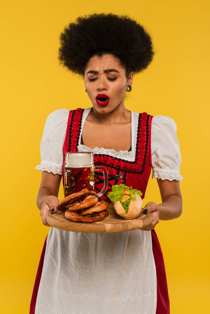 sorprendida camarera bavariana afroamericana con cerveza y deliciosa golosina en bandeja de madera en amarillo - Foto, Imagen