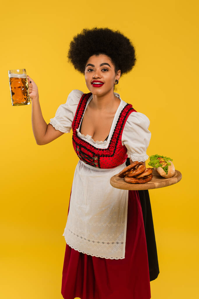 junge afrikanisch-amerikanische Kellnerin im Dirndl serviert Bier und Essen auf Holztablett auf gelb - Foto, Bild