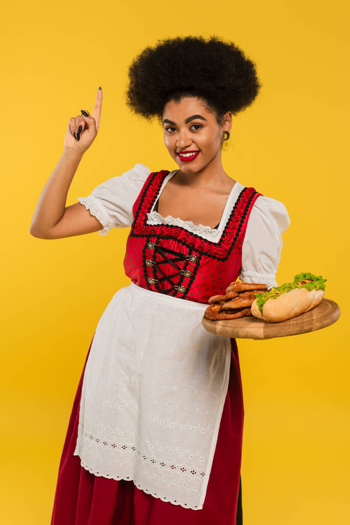 joyful african american oktoberfest waitress with snacks on wooden tray showing idea sign on yellow - Photo, Image