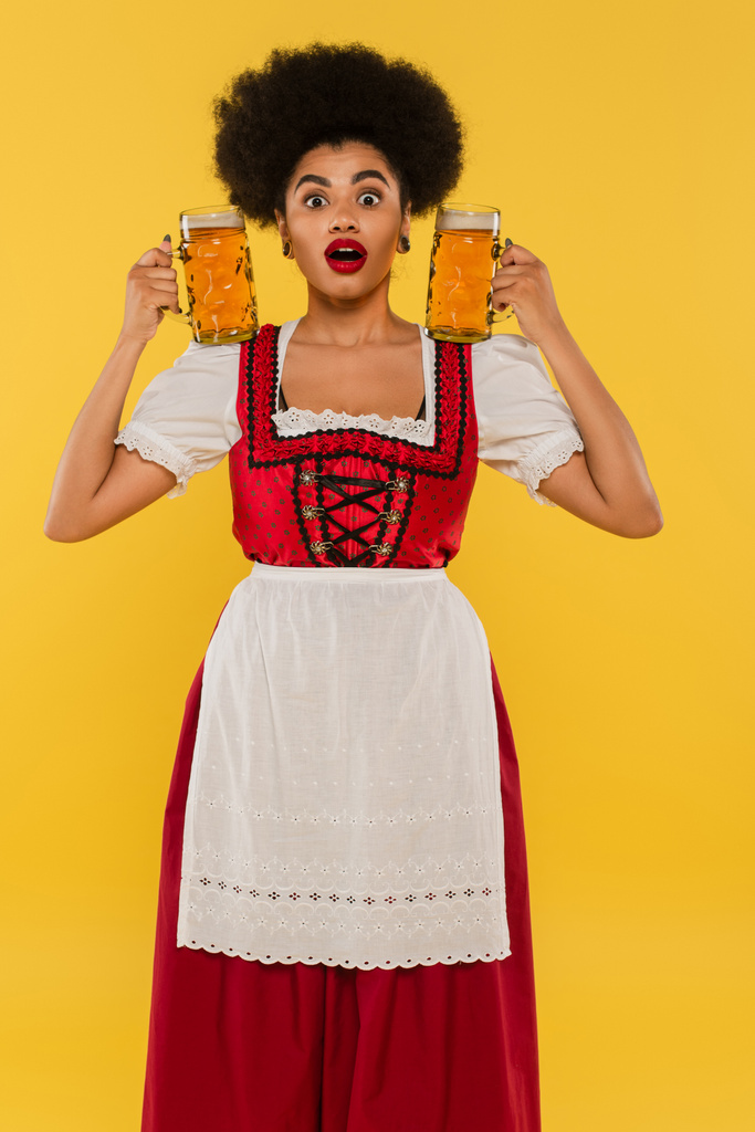 verbaasd afrikaanse Amerikaanse serveerster in traditionele bavariaanse kostuum met biermokken op geel - Foto, afbeelding