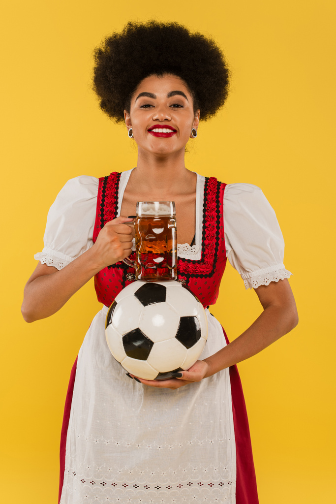 felice cameriera bavarese africana americana con boccale di birra e pallone da calcio sorridente sul giallo - Foto, immagini