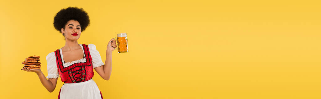 Afrikai amerikai bajor pincérnő Dirndlben sörkorsóval és finom perecekkel a sárga zászlón. - Fotó, kép