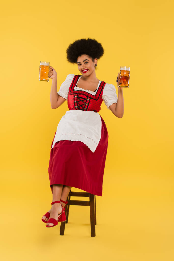 emocionado africano americano bavarian camarera en dirndl sentado en silla con jarras de cerveza en amarillo - Foto, imagen