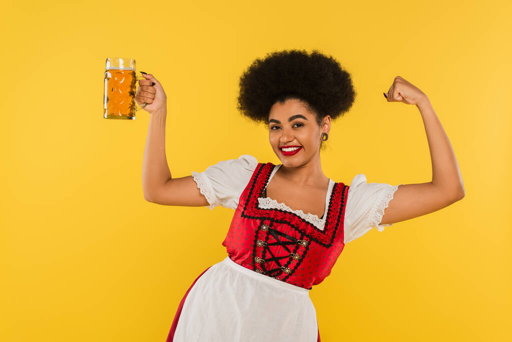 fröhliche afrikanisch-amerikanische Oktoberfest-Kellnerin mit Bierkrug zeigt Triumphgeste auf gelb - Foto, Bild