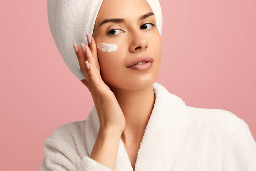Crop giovane modello femminile in accappatoio bianco e asciugamano avvolto intorno alla testa applicando crema idratante sul viso e guardando lontano sullo sfondo rosa - Foto, immagini