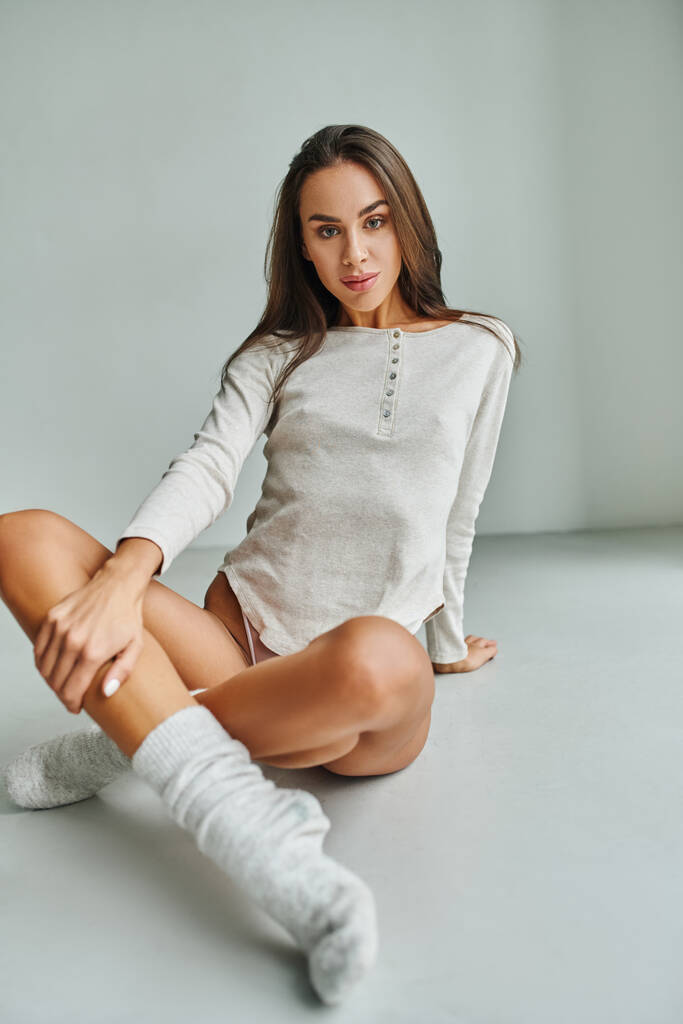 aantrekkelijke jonge vrouw in sokken en shirt met lange mouwen zitten op de vloer thuis en kijken naar de camera - Foto, afbeelding