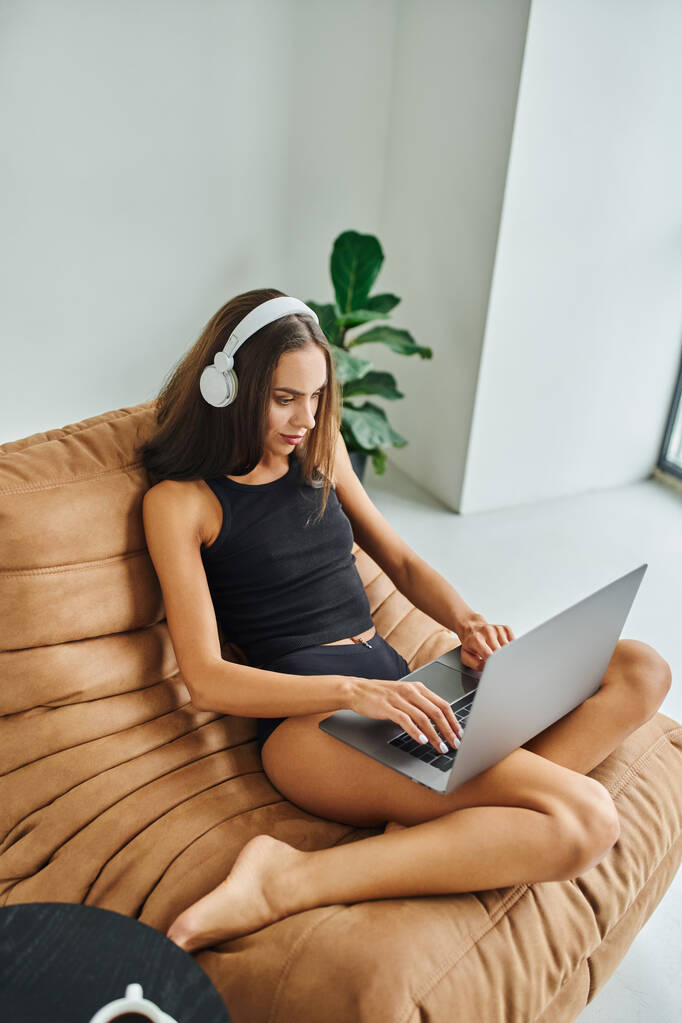 όμορφη freelancer σε ασύρματα ακουστικά χρησιμοποιώντας φορητό υπολογιστή και κάθεται στην καρέκλα τσάντα φασόλι, νεαρή γυναίκα - Φωτογραφία, εικόνα
