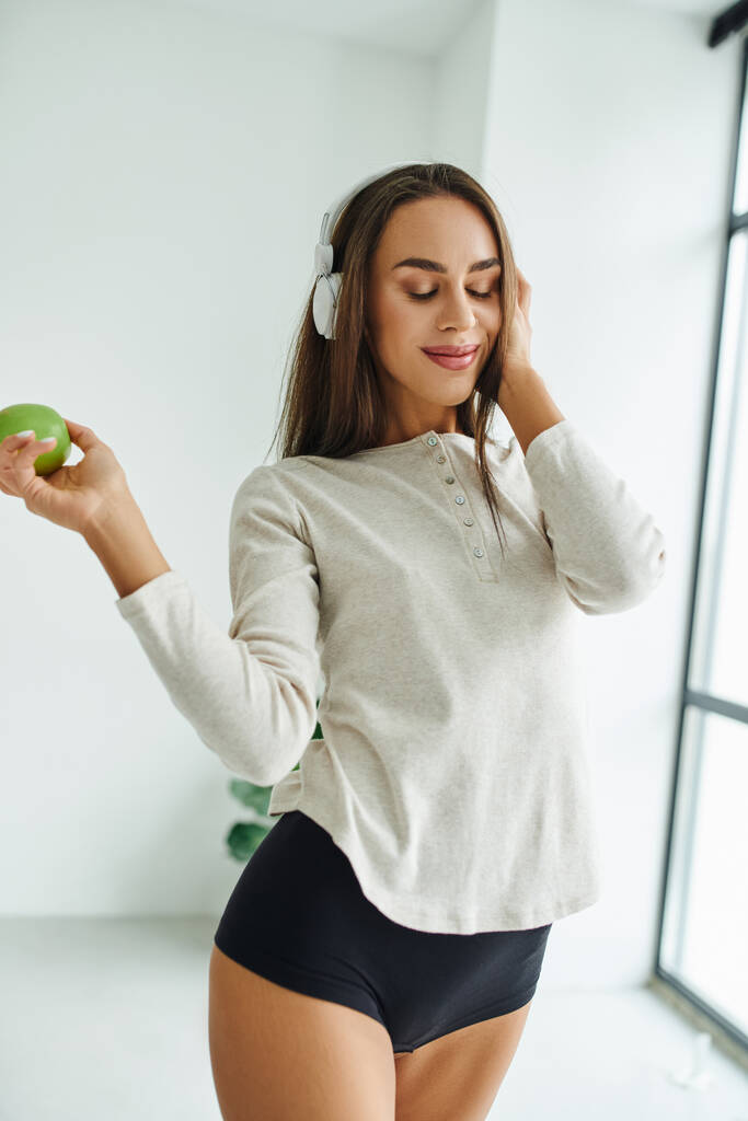 radostná žena v dlouhém rukávu a kalhotky poslouchá hudbu ve sluchátkách a drží jablko - Fotografie, Obrázek