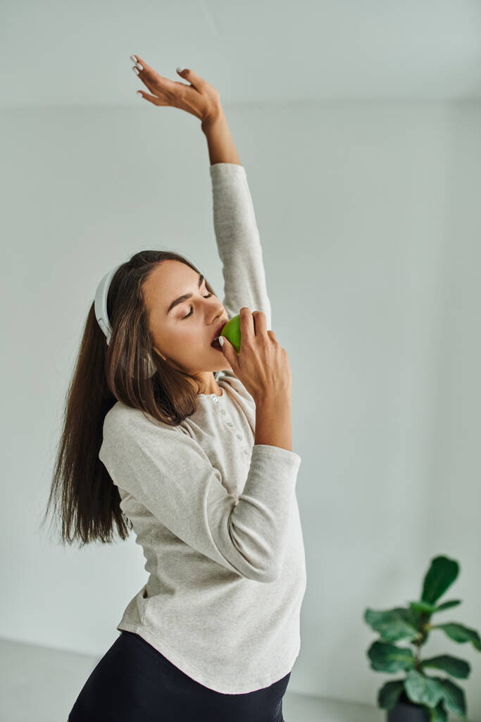 брюнетка женщина в длинном рукаве слушать музыку в наушниках и кусать зеленое яблоко, наслаждаясь вкусом - Фото, изображение