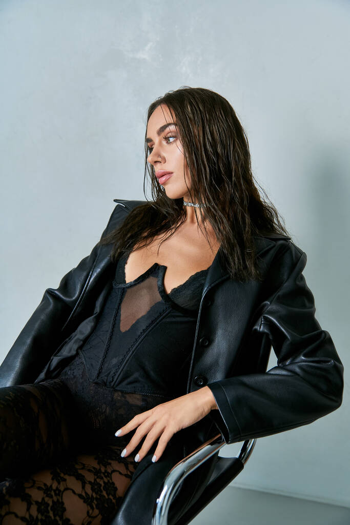 seducente donna con i capelli bagnati indossa cappotto di pelle nera e corsetto mentre seduto sulla sedia - Foto, immagini