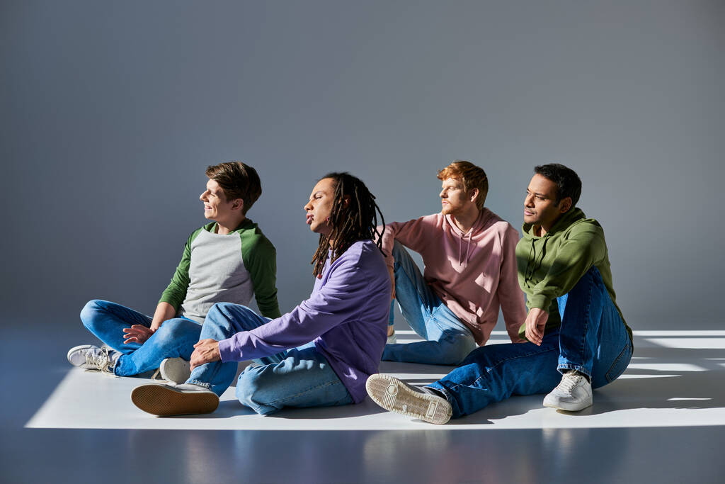four young men in casual outfits sitting on floor on grey background and looking away, diversity - Photo, Image