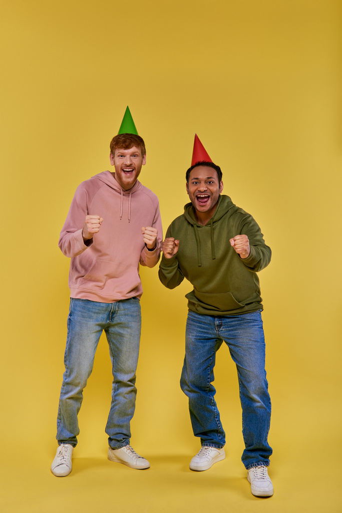 Két jóképű multiracionális férfi szülinapi kalapban, állva és ököllel ünnepelve, születésnap. - Fotó, kép
