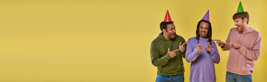 tre uomini multietnici in cappelli di compleanno che sorridono e puntano il dito contro macchina fotografica, compleanno, stendardo - Foto, immagini
