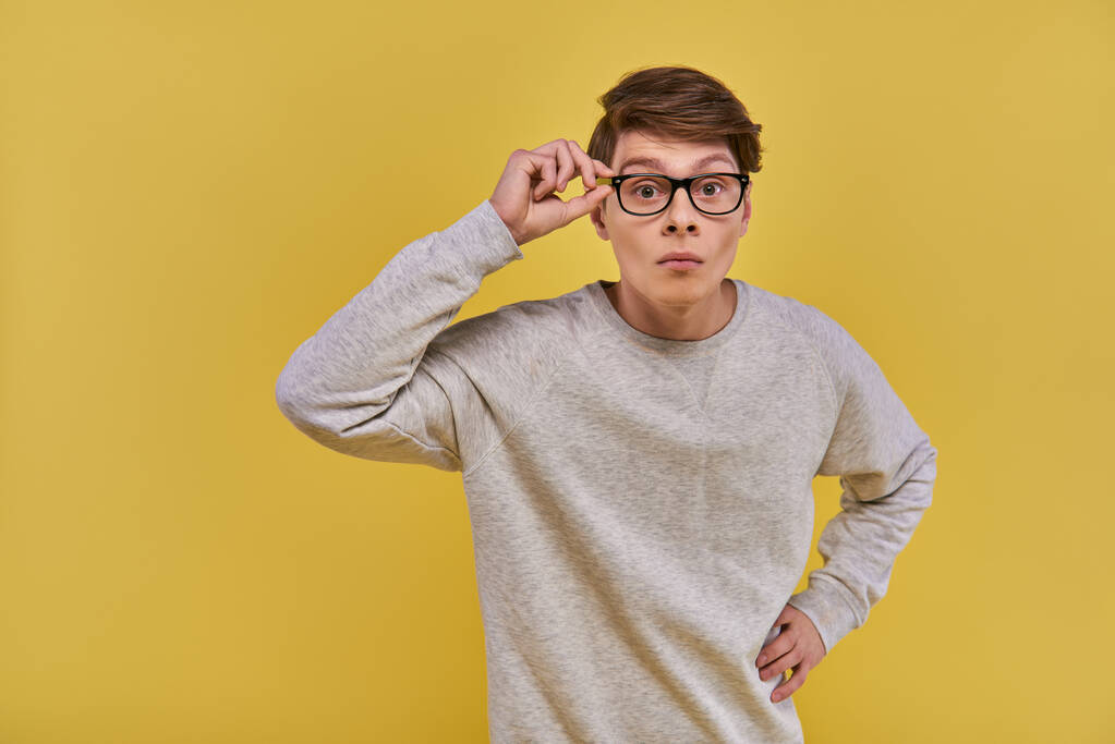 surprised young man in glasses and white casual sweatshirt touching his glasses looking at camera - Photo, Image