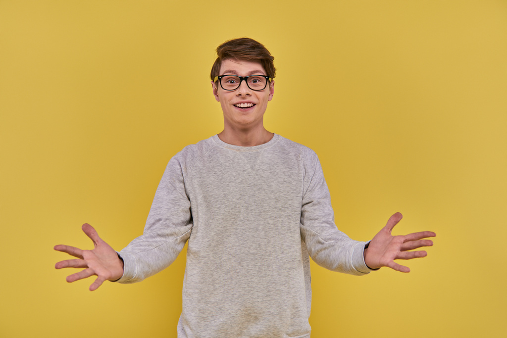 alegre hombre sorprendido en traje casual y gafas levantando las manos con las palmas abiertas sobre fondo amarillo - Foto, imagen