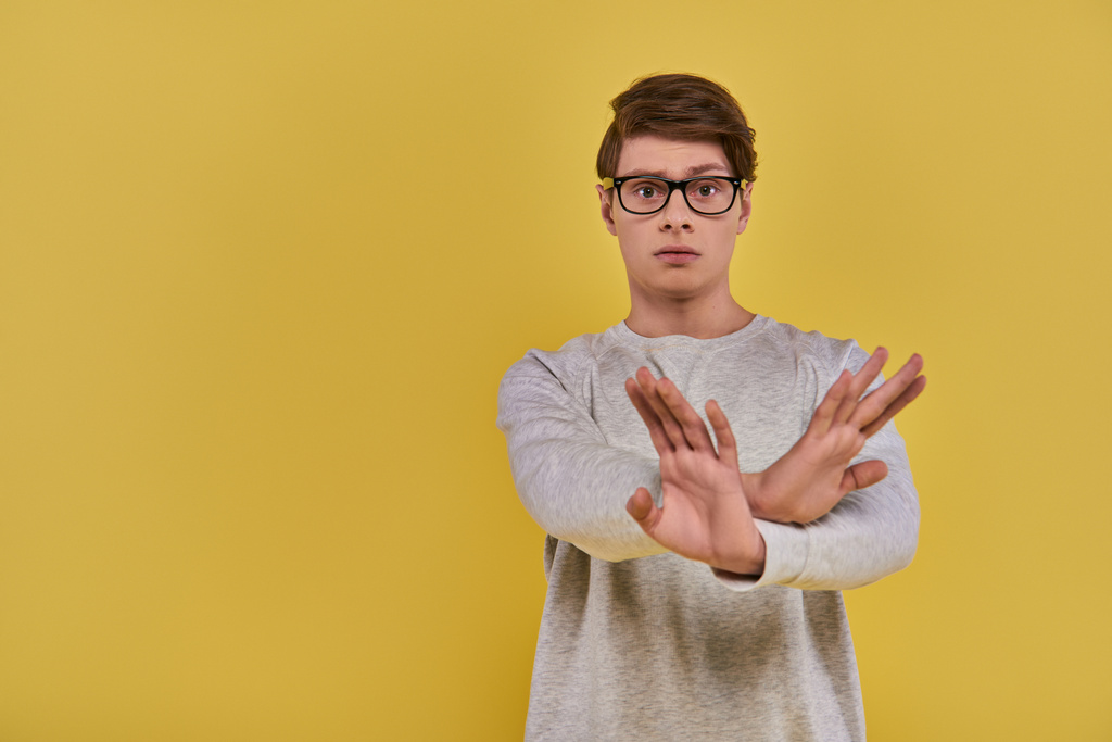 joven hombre triste en sudadera blanca con gafas que muestran gesto cruz negación mano sobre fondo amarillo - Foto, imagen