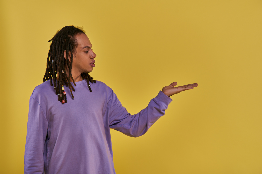 спантеличений афроамериканський чоловік у фіолетовому светрі з дредлоками тримає руку порожньою долонею вгору - Фото, зображення
