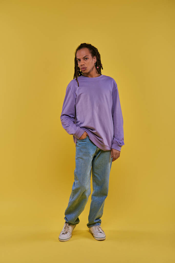 joven afroamericano en sudadera púrpura y jeans con rastas posando sobre fondo amarillo - Foto, imagen