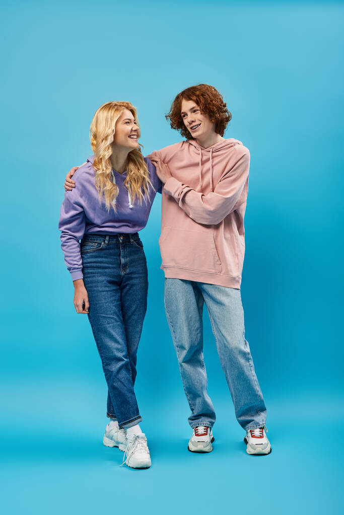 веселые друзья-подростки в толстовках и джинсах, обнимающиеся и смотрящие друг на друга на синем, полном длине - Фото, изображение