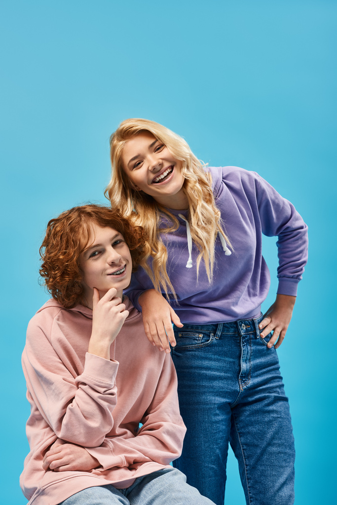 cheerful teenage couple of friends in stylish hoodies posing and smiling at camera on blue - Photo, Image