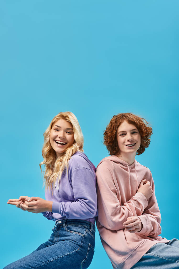 веселые друзья-подростки в модных толстовках сидят спиной к спине и смеются над камерой в студии на голубом - Фото, изображение