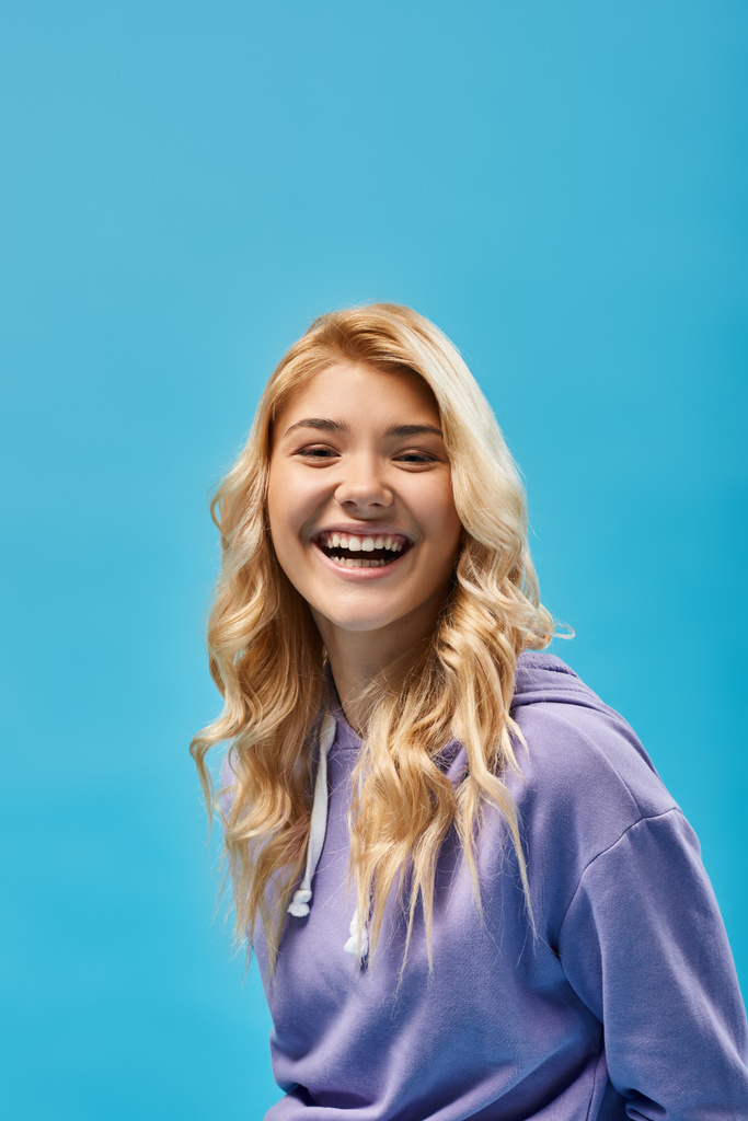 excited blonde teenage girl in hoodie looking at camera and laughing on blue, happiness - Photo, Image
