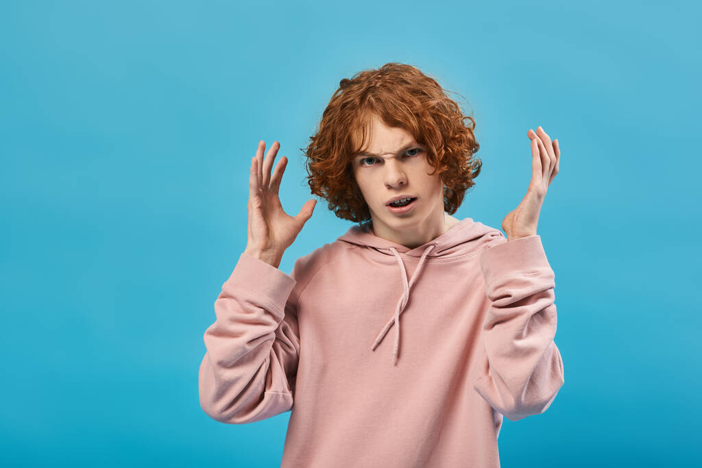 irritierter Teenager mit roten welligen Haaren gestikuliert und blickt in die Kamera auf blauer, schlechter Laune - Foto, Bild