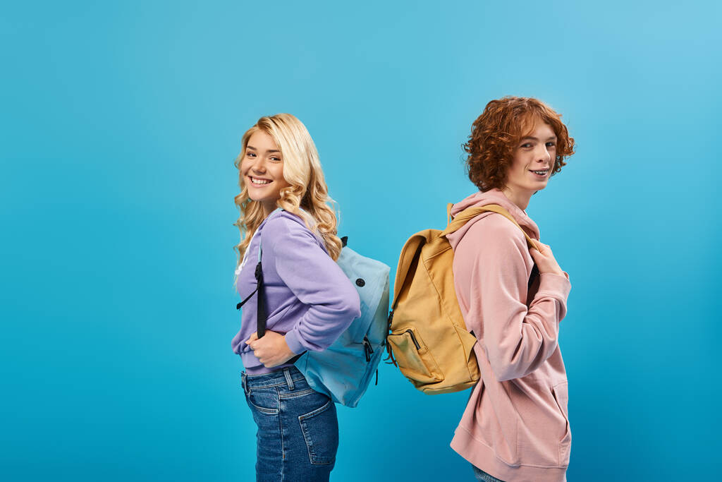счастливые подростки в толстовках, стоящие спиной к спине с рюкзаками и смотрящие в камеру на голубом - Фото, изображение