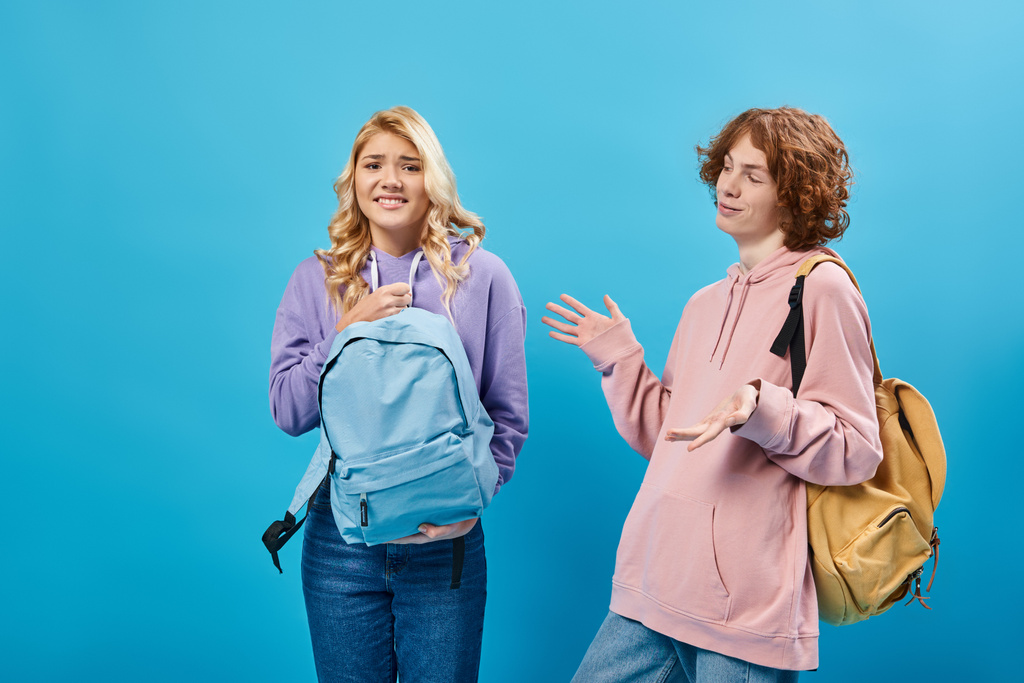 fröhlich rothaarige Teenie-Junge gestikuliert in der Nähe unzufriedene Freundin hält schweren Rucksack auf blau - Foto, Bild