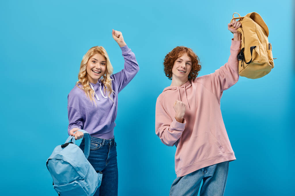 aufgeregte Teenager in trendiger Freizeitkleidung mit Rucksäcken und Erfolgsgeste auf blauem Grund - Foto, Bild