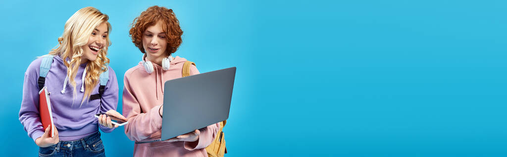 gelukkig tiener meisje met smartphone en notebook op zoek naar laptop in de buurt roodharige vriend op blauw, banner - Foto, afbeelding