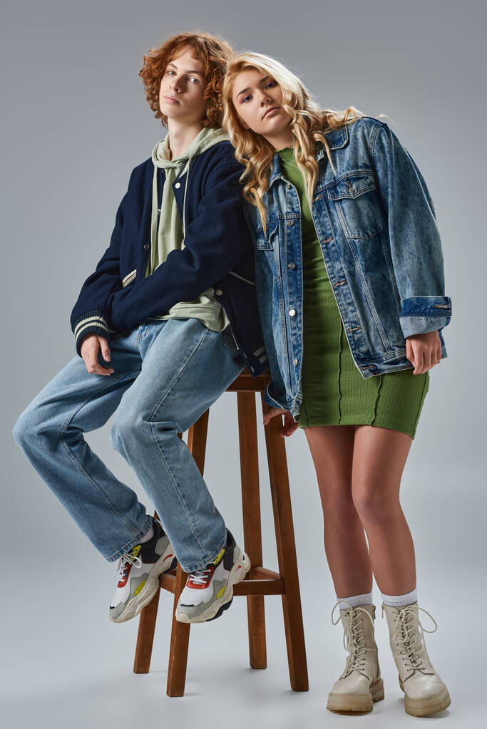 blonde teenage girl leaning on redhead boyfriend sitting on high stool on grey, youth fashion - Photo, Image