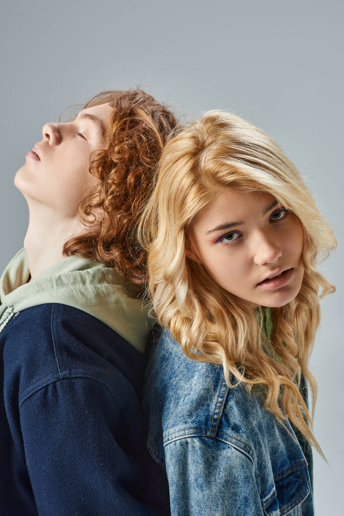 stylish blonde teen girl looking at camera back to back with redhead friend with closed eyes on grey - Photo, Image