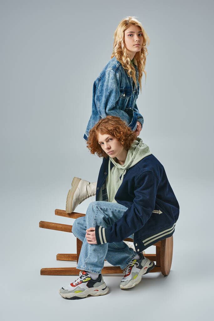 adolescente pareja de amigos en moda ropa casual posando cerca de taburete y mirando a la cámara en gris - Foto, Imagen