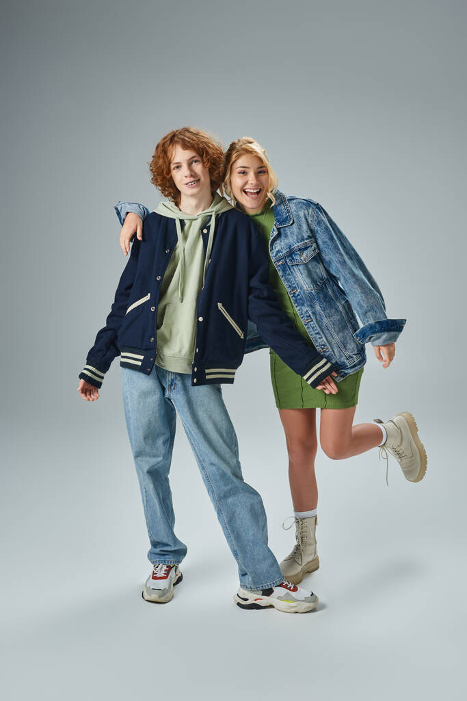 fröhliches Teenie-Paar in modischer Freizeitkleidung, das in grau in voller Länge in die Kamera lächelt - Foto, Bild