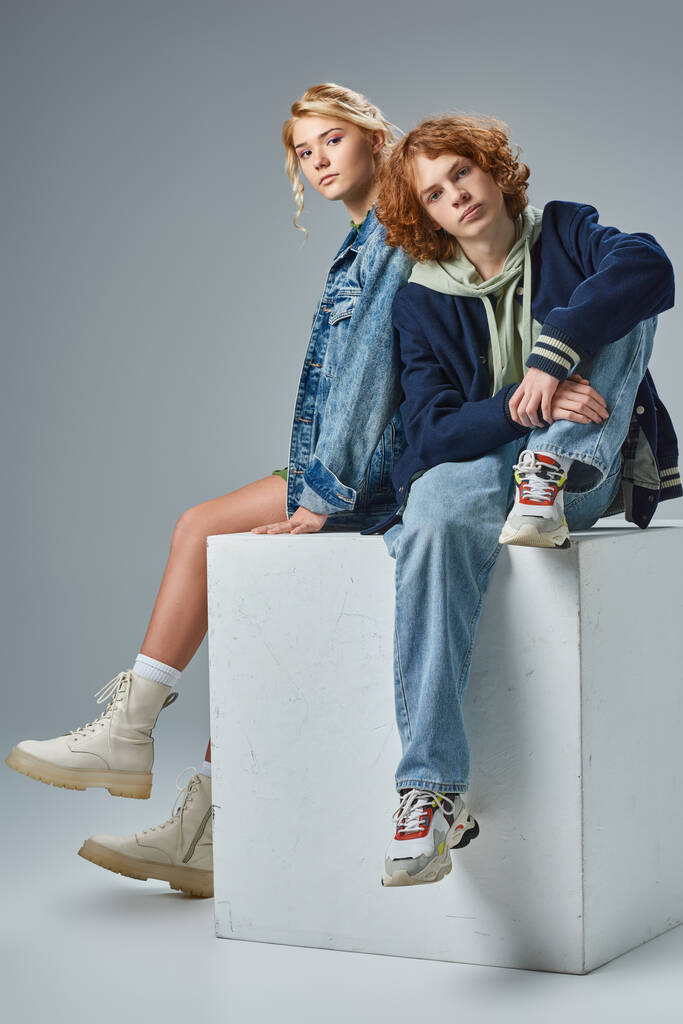 teenage friends in stylish  casual clothes sitting on white cube and looking at camera on grey - Photo, Image
