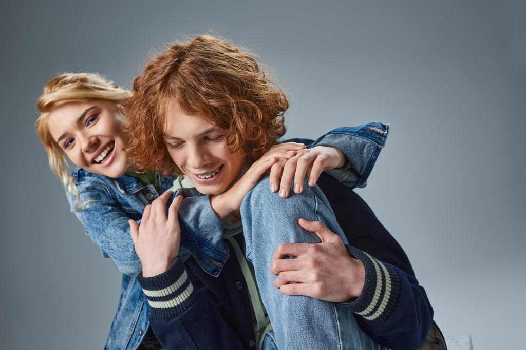 веселая блондинка-подросток обнимает модного рыжеволосого парня и улыбается в камеру на серой - Фото, изображение