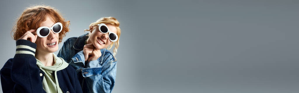 щасливі друзі-підлітки в стильному повсякденному вбранні і сонцезахисних окулярах позують і посміхаються на сірому, банер - Фото, зображення