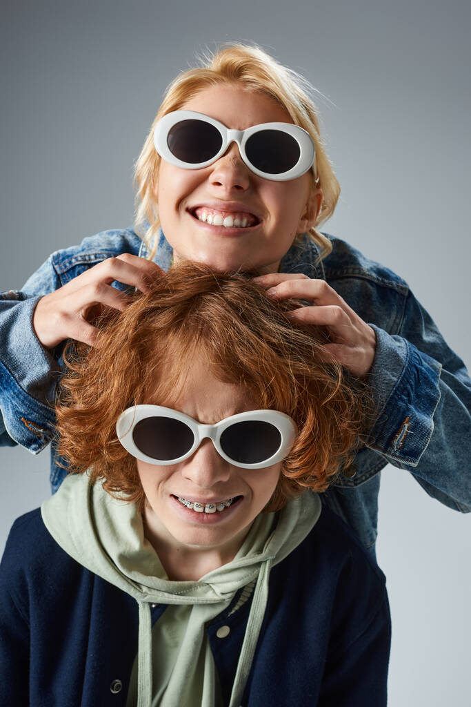 vzrušené blondýny teen dívka v módní sluneční brýle dotýkat zrzavé vlasy přítele na šedé, mládež život - Fotografie, Obrázek