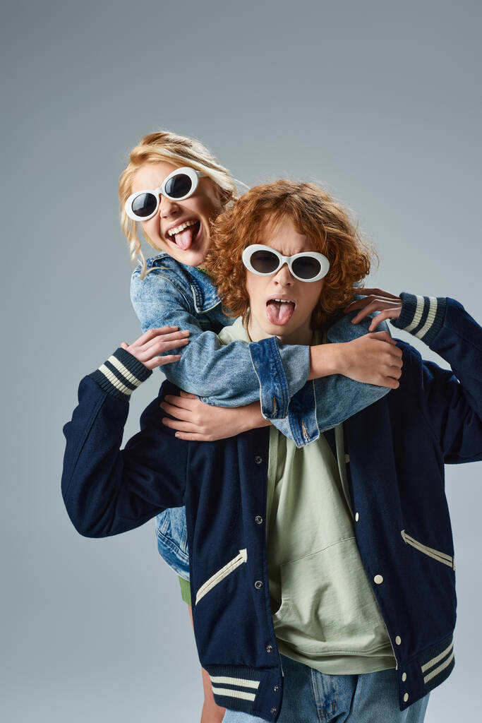 стильные подростки в повседневной одежде и солнцезащитных очках, торчащие языками, веселясь на сером - Фото, изображение