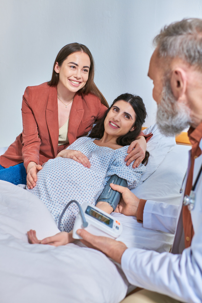 cheerful lesbian couple hugging and looking at doctor that measuring pressure, ivf concept - Photo, Image