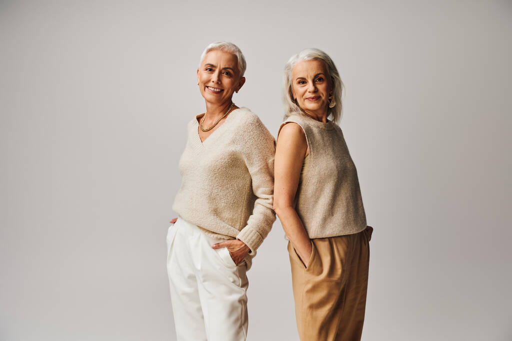 smiling senior women in fashionable clothes standing back to back with hands in pockets on grey - Photo, Image
