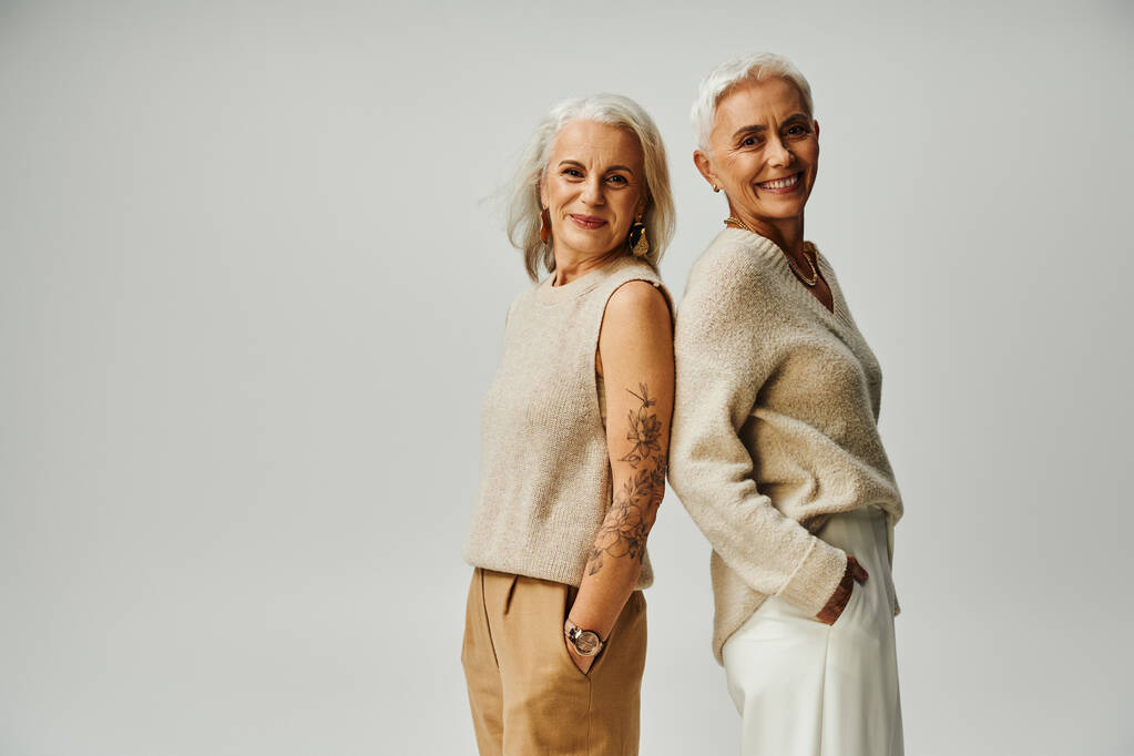 señoras maduras felices mirando a la cámara mientras que de pie espalda con espalda en gris, seniors de moda - Foto, Imagen