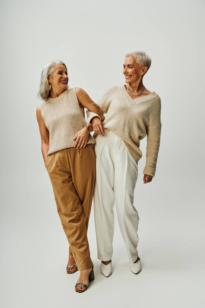 volledige lengte van vrolijke en stijlvolle vrouwelijke vrienden kijken naar elkaar op grijze, senior mode - Foto, afbeelding