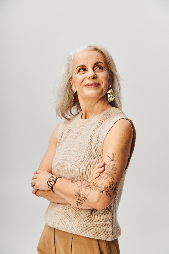 dame tatouée mature en haut tricoté debout avec les bras croisés et regardant loin sur gris, positivité - Photo, image