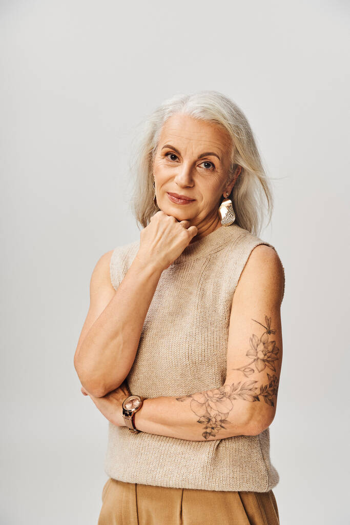 старшая татуированная женщина в пастельном топе держит руку возле подбородка и смотрит в камеру на сером фоне - Фото, изображение