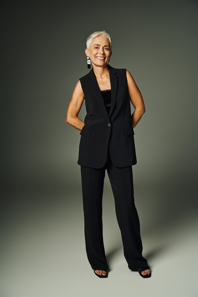 mujer mayor de moda con sonrisa radiante posando en traje elegante negro en gris, longitud completa - Foto, Imagen