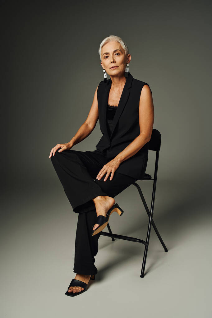 modische Seniorin in schwarzer eleganter Kleidung sitzt auf einem Stuhl auf grau, klassischer Mode und Alter - Foto, Bild